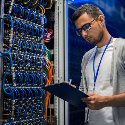 Porträt des modernen jungen Mannes, der einen Laptop hält, der im Serverraum steht und mit einem Computer in blauem Licht arbeitet
