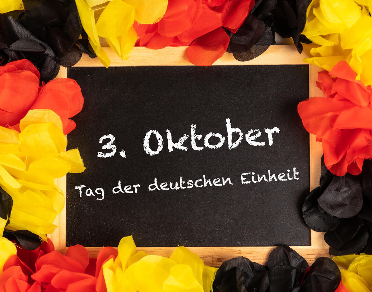 Bild vergrößern: Tafel mit Aufschrift: 3. Oktober Tag der deutschen Einheit, umrahmt mit bunten Blumen (schwarz, rot , gelb)