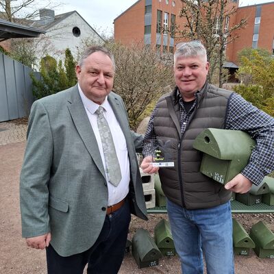 Bild vergr&ouml;&szlig;ern: Zwei M�nner stehen drau�en vor Geb�uden und B�umen. Der Mann links tr�gt Anzug und Krawatte, der Mann rechts eine Weste und Jeans. Sie blicken in die Kamera. Der Mann rechts h�lt mehrere gr�n gestrichene Nisthilfen sowie eine kleine Auszeichnung in der Hand. Hinter ihnen liegen weitere Nisthilfen auf einem Tisch und am Boden. Die Szene zeigt eine offizielle �bergabe von Vogel-Nistk�sten.
