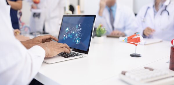Bild vergr&ouml;&szlig;ern: Person im wei�en Kittel nutzt einen Laptop mit medizinischen Symbolen auf dem Bildschirm, w�hrend im Hintergrund medizinisches Personal in einer Besprechung sitzt. Ein Augenmodell steht auf dem Tisch.
